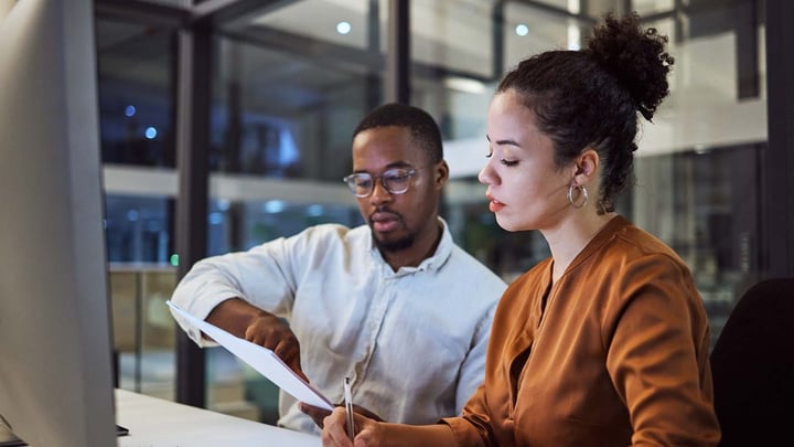 Two colleagues are reviewing and working on a document together at a desk in an office, possibly during evening hours.