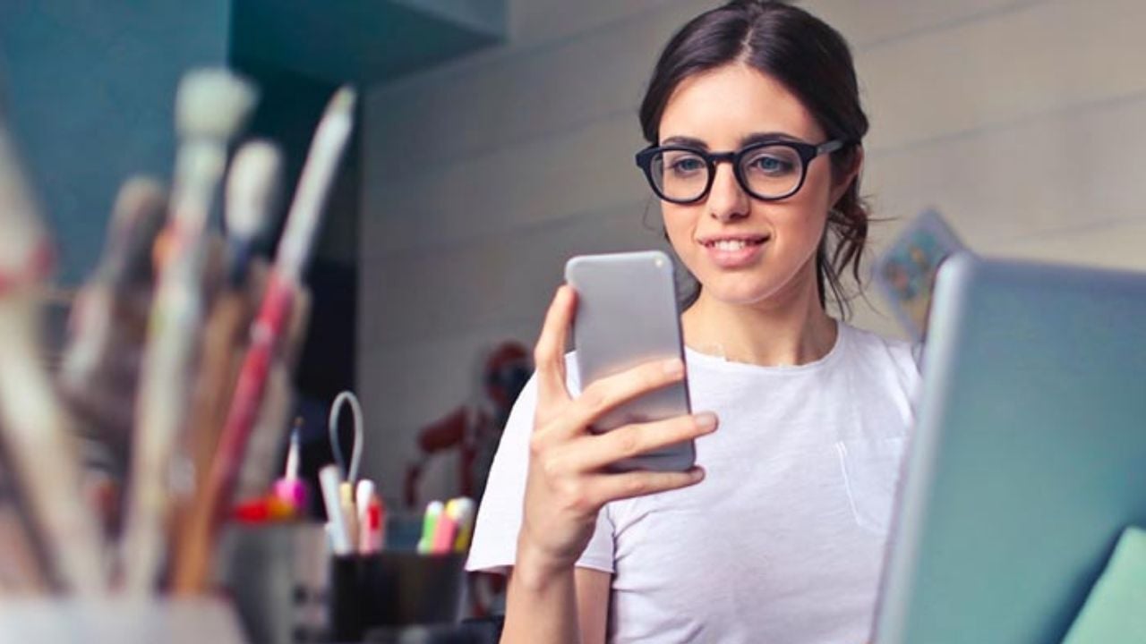 A woman working or browsing on her smartphone while seated at a desk with a laptop and art supplies nearby.