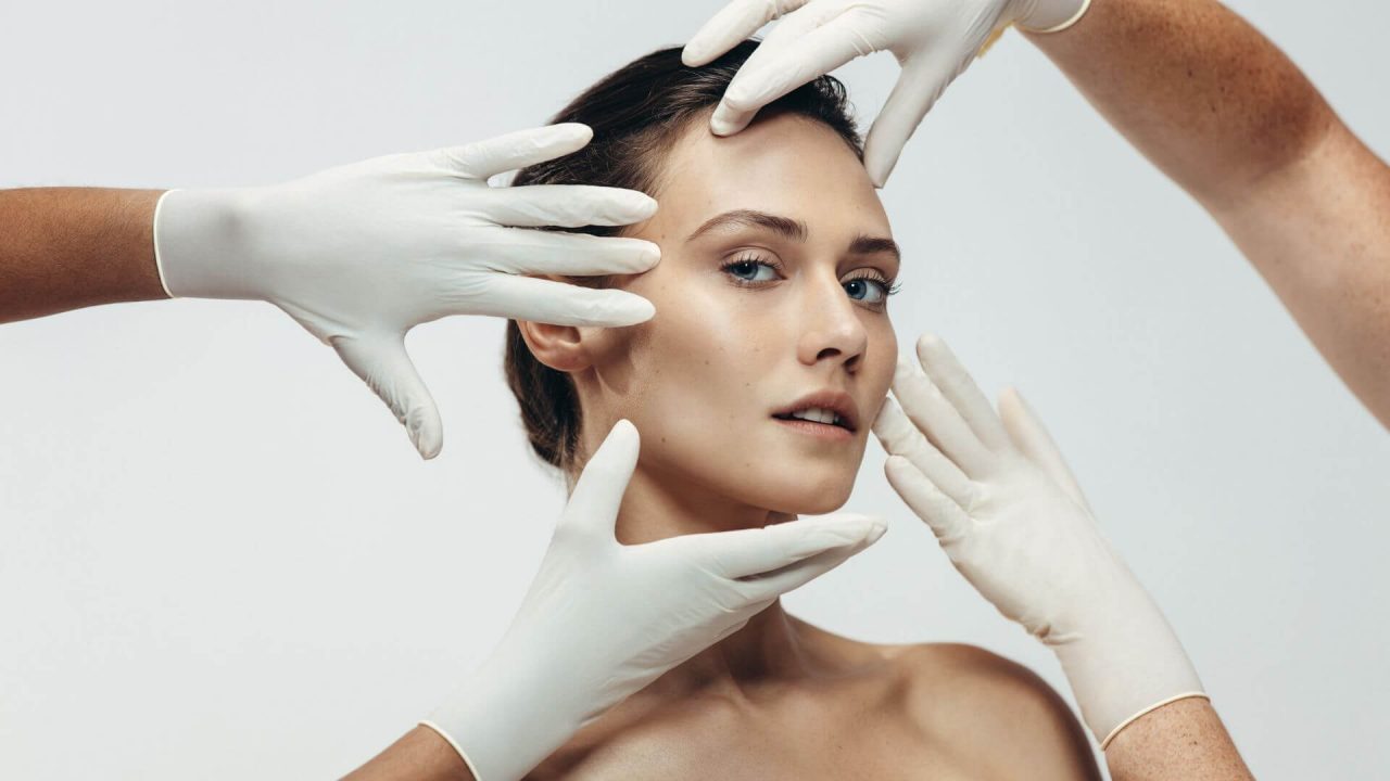 A woman's face being examined by several sets of gloved hands, suggesting a cosmetic procedure consultation or skin assessment.