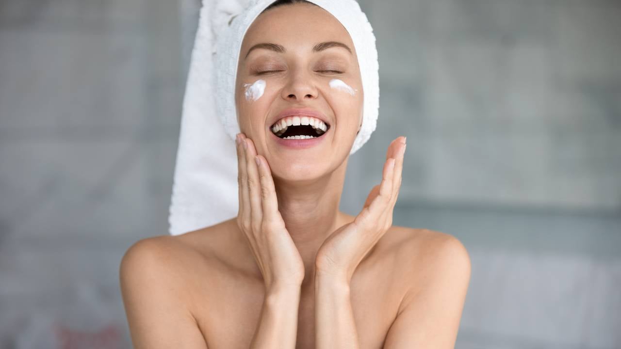 A happy woman enjoying her skincare routine, applying cream to her face after showering, with a towel on her head and a bright smile.