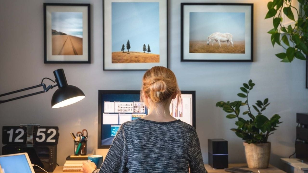 A home office or workspace setup with a woman working on a computer, framed by artwork on the wall, a desk lamp, and a potted plant.